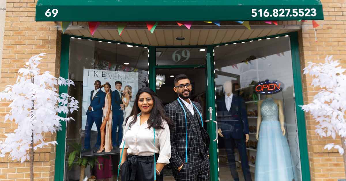 Exterior shot of Fashion Dreams in Syosset NY where one window has a fashion poster and the other window has a manikin of a man in a suit and a woman in a cocktail dress. The owner, Sanu and her son are in front facing the camera and smiling.