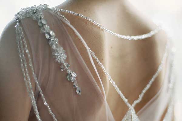 Closeup of the upper part of a woman's back wearing a wedding dress that has hand-sewn beaded edges and a long string of jewels draped over her shoulder.